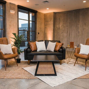 A stylish lounge setup featuring a sleek black vegan leather sofa paired with a cane rattan and black coffee table. The arrangement is complemented by elegant Calvin chairs and layered area rugs that add texture and depth. The overall look is modern and cohesive, with a polished, curated feel. (Note: image is AI-generated.)