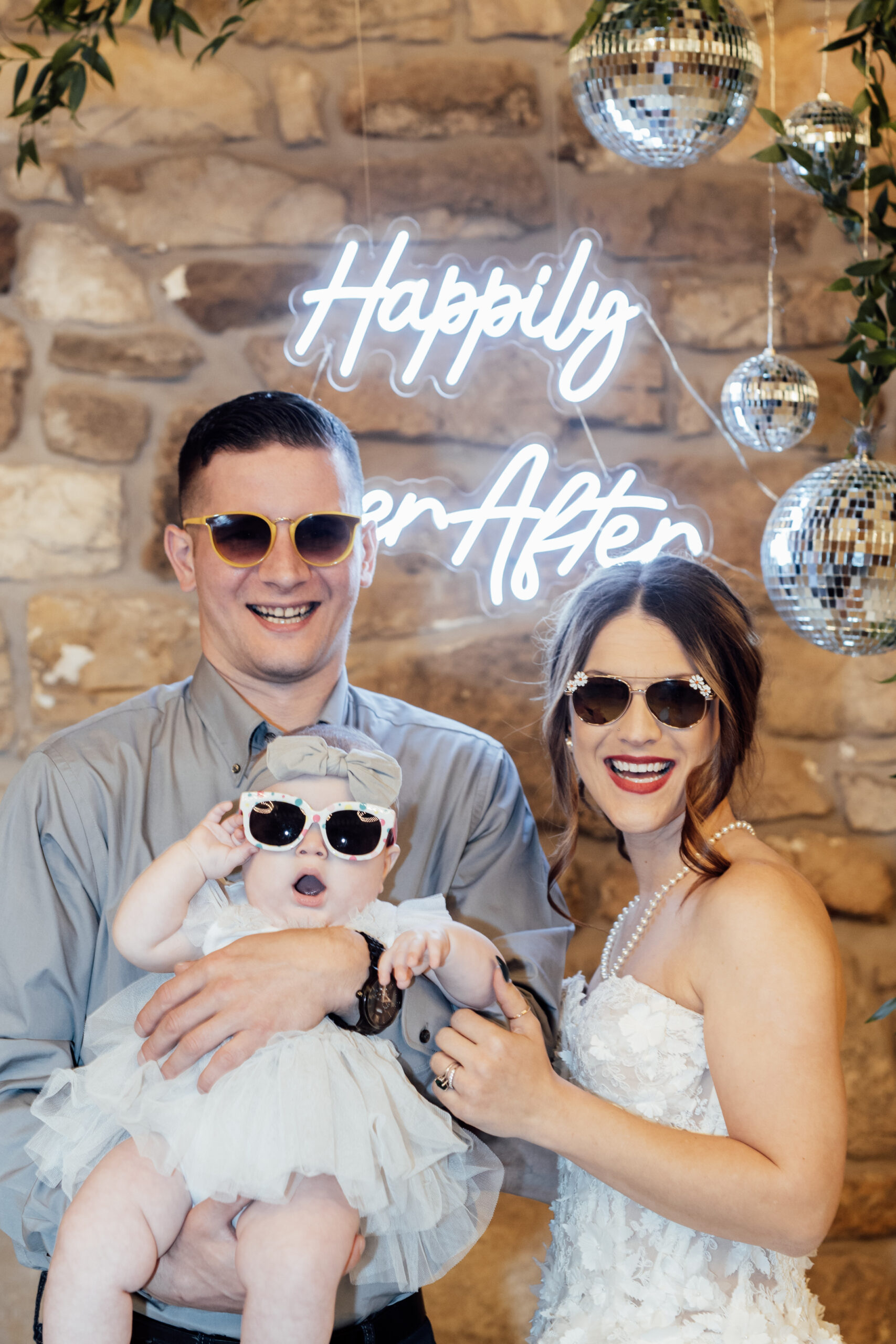 Image of bride, groom, and baby wearing sunglasses in front of "happily Ever After" neon sign