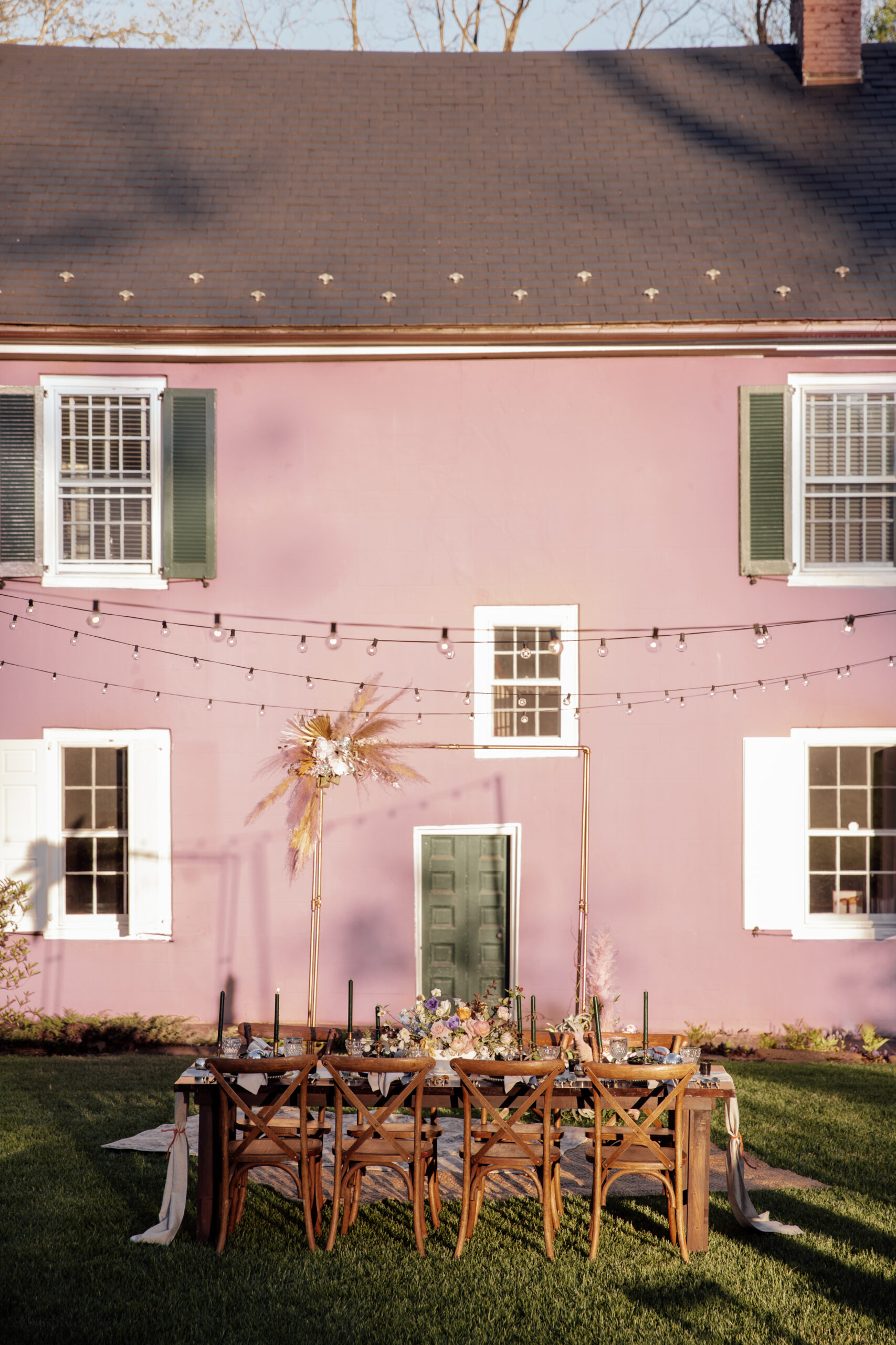 Image of farmhouse tables, crossback chairs, and copper wedding arch set up in courtyard with pink house in the background