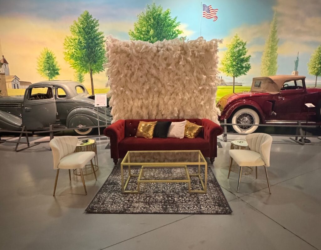 Feather wall with red sofa and 2 ivory accent chairs with black rug, gold coffee table, 2 gold accent tables with a variety of pillows
