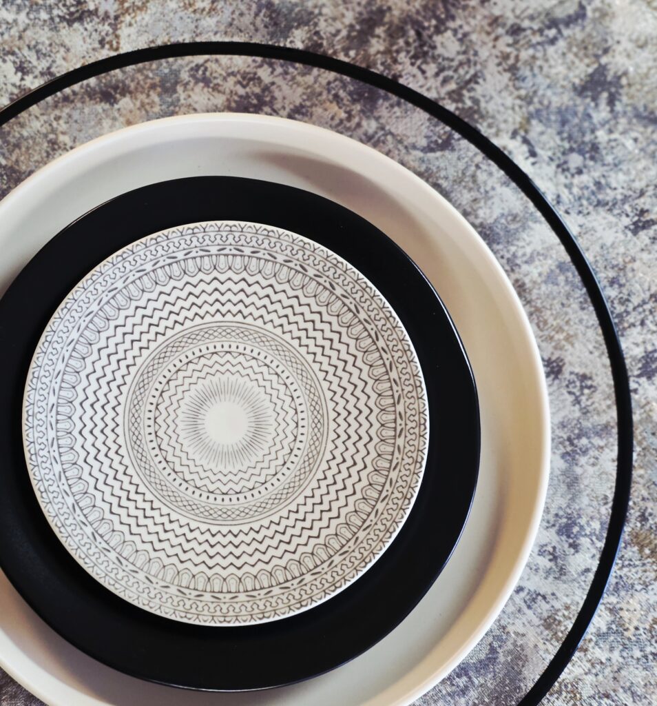 Black glass rim charger with sand stoneware dinner plate, black stoneware coupe salad plate, and cream and black accented b&b plate. All on a variety of brown-hued marbled linen.